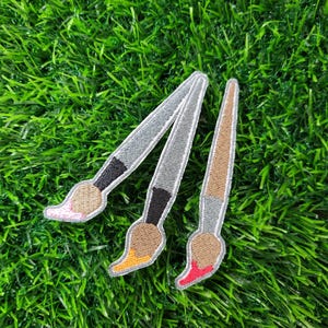 May include: Three embroidered patches of paintbrushes with gray handles, black ferrules, and colorful bristles. The bristles are pink, brown, and red, with yellow and orange accents. The patches are on a green, grassy surface.