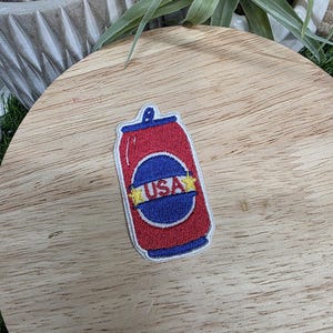 Pu&ograve; includere: Una toppa rossa, bianca e blu a forma di lattina di soda. La toppa presenta la scritta "USA" in un cerchio blu con stelle gialle. La lattina &egrave; rossa con la parte superiore e inferiore blu.