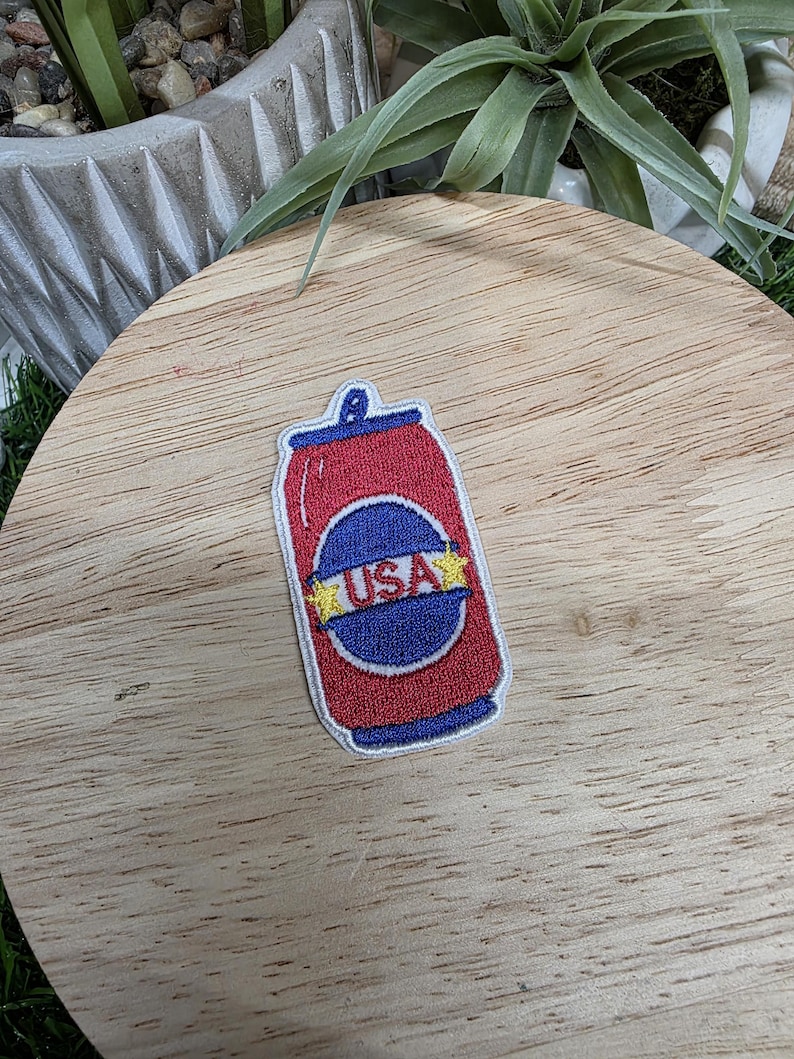 Pu&ograve; includere: Una toppa rossa, bianca e blu a forma di lattina di soda. La toppa presenta la scritta "USA" in un cerchio blu con stelle gialle. La lattina &egrave; rossa con la parte superiore e inferiore blu.