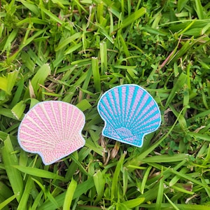 May include: Two embroidered fabric patches shaped like seashells. One is pink and the other is blue with a purple stripe.