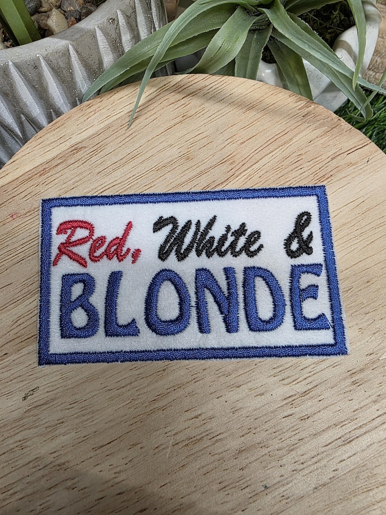 Pu&ograve; includere: Toppa ricamata con le parole "Red, White & BLONDE" in rosso, nero e blu. La toppa ha uno sfondo bianco con un bordo blu. Il testo &egrave; un mix di lettere corsive e a blocchi.