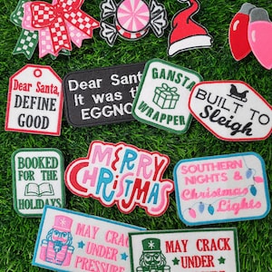 May include: A collection of Christmas-themed patches with various designs and text. Includes a red and white bow, candy, Santa hat, Christmas lights, and patches with phrases like "Dear Santa, Define Good" and "Merry Christmas."