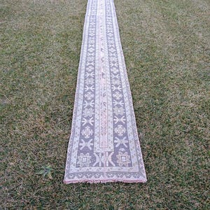 Peut inclure: Un long tapis rectangulaire avec un motif détaillé de formes géométriques et de motifs floraux dans des tons de gris, de blanc et de rose. Le tapis est posé sur de l'herbe verte, mettant en valeur son design et sa texture complexes.