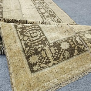 Könnte beinhalten: Beige-brauner Vintage-Teppich mit geometrischem Bordürendesign. Der Teppich hat ein abgenutztes Aussehen mit sichtbaren Gebrauchsspuren und einem Fransenrand. Der Teppich ist gefaltet und zeigt das Muster und die Textur.