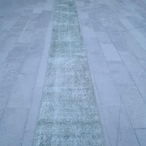 May include: A rectangular, faded green rug with a distressed appearance. The rug is centered on a gray stone surface, with a long, narrow design. The rug's texture appears soft, with a vintage aesthetic.