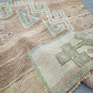 May include: A hand-knotted wool rug featuring a textured pile in shades of beige and tan. The surface displays a geometric pattern in muted green and cream, with a fringed edge visible along the bottom corner of the folded textile.