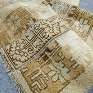 May include: A beige and brown vintage rug with geometric patterns. The rug features a patchwork design with various shades of beige and brown, creating a warm and inviting aesthetic. The rug's edges are frayed, adding to its antique charm.