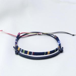 May include: A dual-layered bracelet featuring one band with a dark blue base and multi-coloured woven stripes in yellow, green, and red, paired with a second solid dark blue cord band. Both bands are connected by adjustable sliding knots.