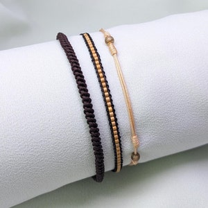 May include: Three bracelets stacked on a white surface. The top bracelet is a thin beige cord with a small gold bead. The middle bracelet is a brown cord with small beige beads. The bottom bracelet is a thick brown cord.