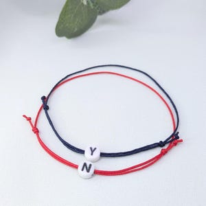 May include: Two friendship bracelets with the letters "N" and "Y" on white beads. The bracelets are made with red and blue string.