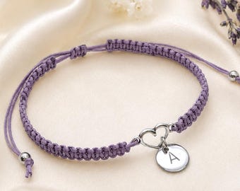 May include: A purple cord bracelet featuring a silver-toned heart-shaped charm and a small circular pendant engraved with the letter A. The bracelet has a braided design with adjustable sliding knots and small silver-toned beads at the ends.