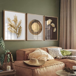 Op de afbeelding: Drie ingelijste gouden bladdrukken op een groene muur boven een bruine leren bank met kussens en een deken. De prints tonen een gestileerd blad, een cirkel met een gouden middelpunt en een gestileerde bloem.