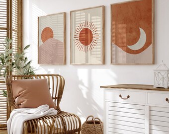 May include: A white cabinet with two drawers and two doors with slatted panels. The cabinet has a wooden top and brass handles. A wicker basket sits on the floor in front of the cabinet. Three framed prints with abstract sun and moon designs hang on the wall behind the cabinet. A wicker chair with a brown cushion sits in front of the cabinet.