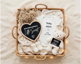 May include: A wicker basket holds baby announcement items. A white onesie with "Baby Adam" and "DUE JULY 2026" is next to a teddy bear holding an ultrasound image. A heart-shaped sign reads "WE'VE BEEN KEEPING A Little Secret".