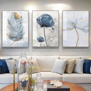 Op de afbeelding: Drie ingelijste wandkunstprints met een blauw en gouden kleurenschema. De linker print toont een enkele blauwe en gouden veer. De middelste print toont twee blauwe bloemen met gouden stelen. De rechter print toont een enkele blauwe en gouden bloem.