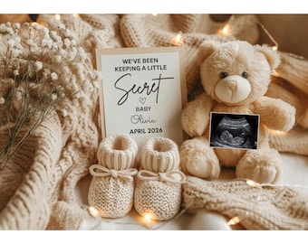May include: A beige teddy bear, baby booties, and a card with the text "We've been keeping a little secret" are arranged on a knitted blanket. The card also says "Baby Olivia April 2026". An ultrasound image is attached to the teddy bear.