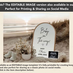 Peut inclure: Un panneau en bois avec le texte "Baby on The way!" et un symbole de c&oelig;ur. Le panneau comprend le nom "Olivia" et la date "AVRIL 2026". L'image pr&eacute;sente &eacute;galement des chaussons de b&eacute;b&eacute;, des fleurs et une image d'&eacute;chographie, le tout sur fond de plage.
