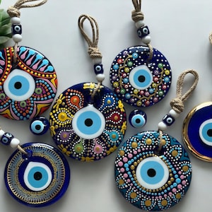 May include: Six blue and white evil eye wall hangings with colourful patterns and gold accents. The evil eyes are round and have a white iris and black pupil. The hangings are attached to twine with white beads.