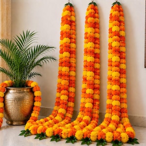May include: Three long, vertical garlands made of alternating bright yellow and vibrant orange marigold flowers, hanging down to the floor. A metallic, hammered-texture gold pot contains a green palm plant, draped with a matching marigold garland.