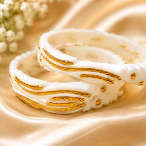 Puede incluir: Dos brazaletes blancos con detalles dorados en relieve y texturizados. Los brazaletes tienen un acabado liso y brillante con intrincados patrones dorados fluidos que envuelven la superficie exterior de cada accesorio circular.