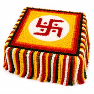 Può includere: Un cuscino quadrato testurizzato con un cerchio bianco centrale contenente un simbolo svastica rosso, circondato da bordi gialli, arancioni e verde scuro. I lati sono decorati con frange verticali multicolori in rosso, giallo, bianco e verde.