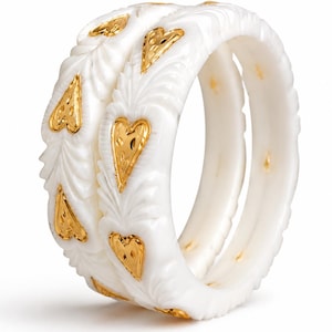 Op de afbeelding: Twee witte armbanden met een uitgesneden bladvormige textuur en ingelegde goudkleurige hartvormen. De armbanden hebben een glad, afgerond profiel met een herhalend patroon van gouden harten die in het witte materiaal zijn gezet.