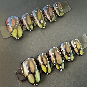 May include: Two sets of press-on nails with a butterfly wing design. The nails are black with iridescent green, gold, and purple accents. Each nail features white polka dots. The nails are displayed on a black surface.