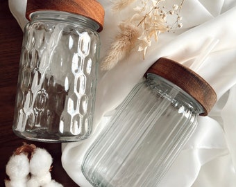 May include: Two clear glass jars with wooden lids. One jar has a textured honeycomb pattern, the other has vertical ridges. The jars are on a white cloth with dried flowers and cotton.