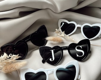 May include: Three pairs of heart-shaped sunglasses with dark lenses. One pair is white, one is black, and one is black with the white letters J and S printed on the lenses. The sunglasses are arranged on a draped, light beige fabric surface.