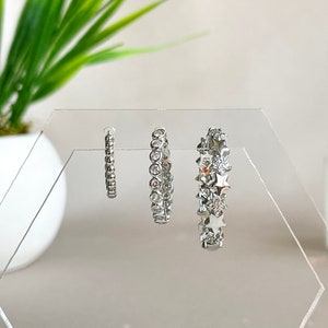 May include: Three pairs of silver hoop earrings displayed on a clear acrylic stand. The earrings vary in design, including small beaded hoops, hoops with clear stones, and hoops with star-shaped embellishments.