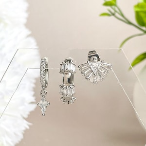 May include: A set of three silver earrings with clear cubic zirconia stones. The earrings are displayed on a white background.