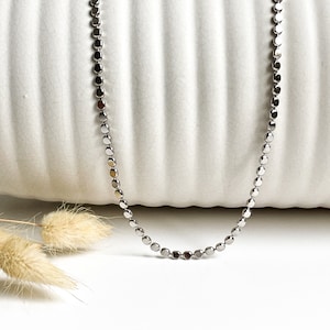 Puede incluir: Un collar de cadena de plata con pequeños eslabones redondos. La cadena está sobre una superficie blanca.