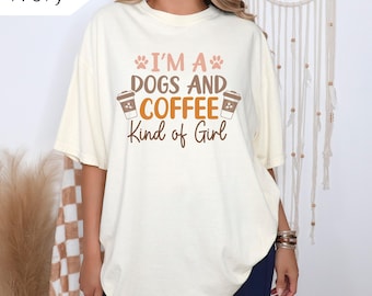 Pode incluir: Camiseta cor marfim com o texto "I'M A DOGS AND COFFEE Kind of Girl" em marrom e laranja. A camiseta apresenta pegadas de patas e gráficos de xícaras de café. A camiseta tem um ajuste folgado.