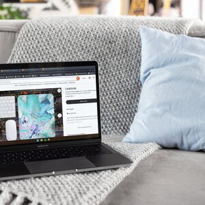 Puede incluir: Un ordenador portátil en un sofá gris con un cojín azul. La pantalla del portátil muestra una lista de productos de Etsy para un teclado y una alfombrilla de ratón personalizados con un diseño abstracto azul y morado. El título de la lista de productos es "CA$29,86 Alfombrilla de escritorio floral personalizada, una alfombrilla de escritorio grande de alta calidad. Sublime añade un toque de clase a tu espacio de trabajo."