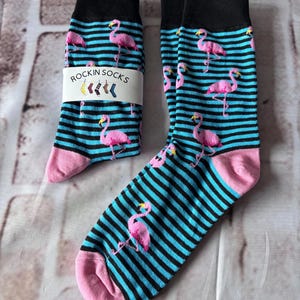 May include: A pair of crew socks featuring a pattern of pink flamingos on a background of horizontal black and bright blue stripes. The socks have solid black cuffs and pink toes. A white paper band labelled ROCKIN SOCKS is wrapped around one sock.