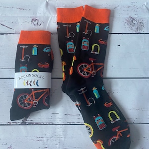 May include: A pair of black socks with an orange cuff. The socks feature a colourful design of bicycles, water bottles, helmets, and other cycling accessories. The socks have the text "Rockin' Socks" on the side.