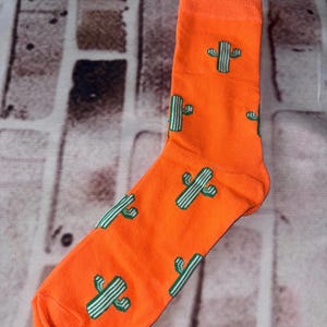 May include: A bright orange crew sock featuring a repeating pattern of small, green, pixelated cactus icons. The sock has a ribbed cuff at the top and a reinforced heel and toe area, showcasing a consistent cactus print across the entire fabric surface.