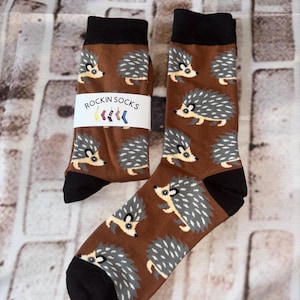 May include: Brown socks featuring a repeating pattern of grey hedgehogs. The socks have black heels, toes, and a black band at the top. The label on the socks reads "ROCKIN SOCKS".
