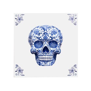 Op de afbeelding: Een decoratieve keramische schedel met een blauw en wit bloemenpatroon. De schedel is gecentreerd op een witte vierkante tegel, met bloemenaccenten in de hoeken. Het ontwerp doet denken aan Delfts blauw aardewerk.