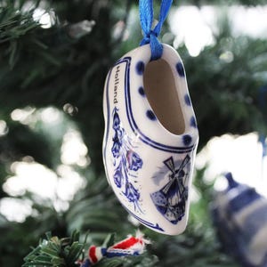 Könnte beinhalten: Ein blauer und weißer Keramik-Klompen-Anhänger mit Windmühle und Blumenmuster. Der Anhänger ist mit "Holland" beschriftet und hängt an einem blauen Band an einem Weihnachtsbaum.