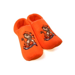 Puede incluir: Un par de pantuflas de felpa naranja con un león y la palabra "Holanda" bordada.