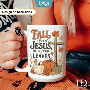 May include: A white ceramic mug featuring a graphic with the text Fall for JESUS, HE NEVER LEAVES. The design includes a wooden cross, three pumpkins in orange, white, and green, and scattered autumn leaves in shades of orange and yellow.