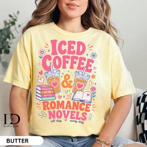 May include: A pale yellow t-shirt featuring a graphic print with the text ICED COFFEE & ROMANCE NOVELS all day every day. The design includes two iced coffee cups, a stack of books, and an open book, decorated with small hearts and flowers.