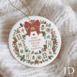May include: A round white ceramic ornament featuring a festive wreath design made of green pine branches, red berries, and pinecones. The wreath is decorated with mahjong tiles, a red bow, and gold bells. The names Emily, Stephanie, Taylor, Stacy, and Katelyn are printed in red text in the centre.