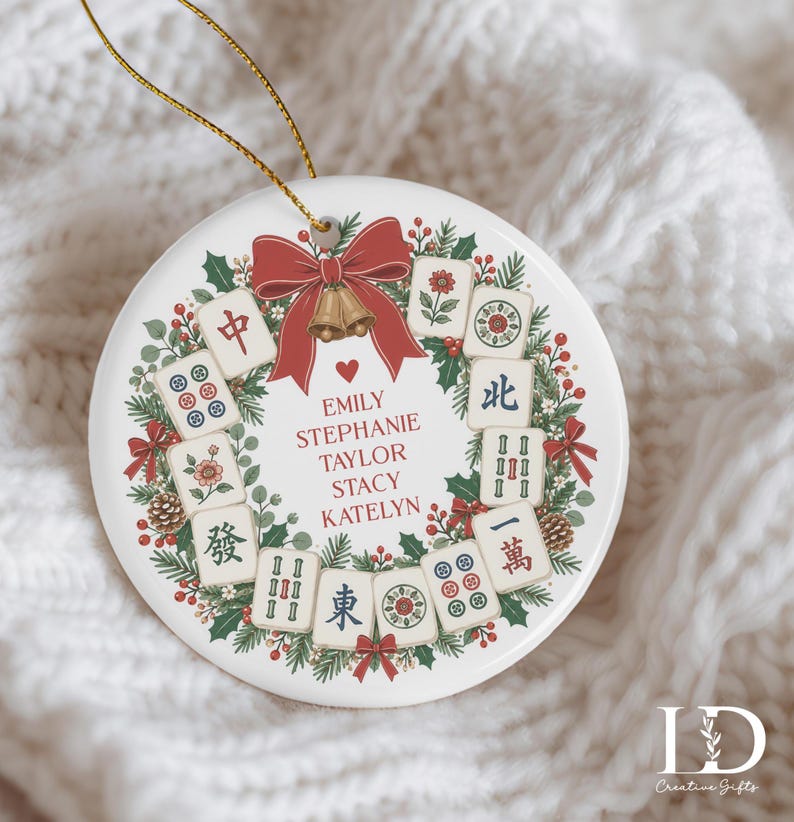 May include: A round white ceramic ornament featuring a festive wreath design made of green pine branches, red berries, and pinecones. The wreath is decorated with mahjong tiles, a red bow, and gold bells. The names Emily, Stephanie, Taylor, Stacy, and Katelyn are printed in red text in the centre.