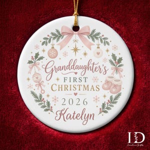 May include: A round white ceramic ornament featuring a pink bow, holly, and baby-themed illustrations. The text reads Granddaughter's FIRST CHRISTMAS 2026 Katelyn. The ornament is suspended by a gold string and decorated with stars and snowflakes.