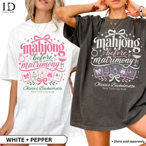 May include: Two oversized t-shirts in white and pepper grey featuring the text 'mahjong before matrimony', 'Olivia's Bachelorette', and 'New York City 2026'. The graphic includes illustrations of mahjong tiles, a ring, a cake, and a bow.