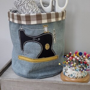 May include: A blue fabric storage bin with a brown and white checked top. The bin has an embroidered image of a black sewing machine with a spool of thread. The bin is filled with white twine and a pair of white scissors. A pincushion with colourful pins is sitting next to the bin.