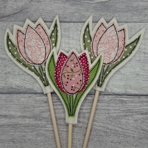 May include: Three decorative tulip flower cutouts on wooden sticks. The tulips have pink and red patterned fabric petals with green leaves and white polka dot accents. The background is a light grey wood grain.