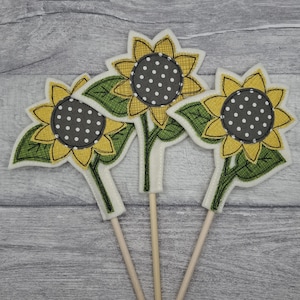 May include: Three felt sunflower decorations with yellow petals, green leaves, and grey polka-dot centres. Each flower is attached to a wooden stick. The sunflowers are arranged against a grey wooden background.